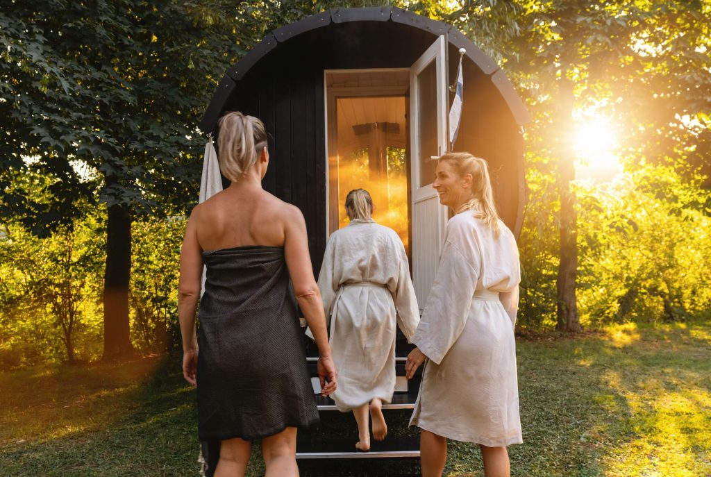 Drei Frauen in Saunamänteln auf dem Weg zum schwarzen Saunafass inmitten der Natur bei Sonnenuntergang. Mobile Sauna zur Miete oder zum Verkauf in Regensburg.